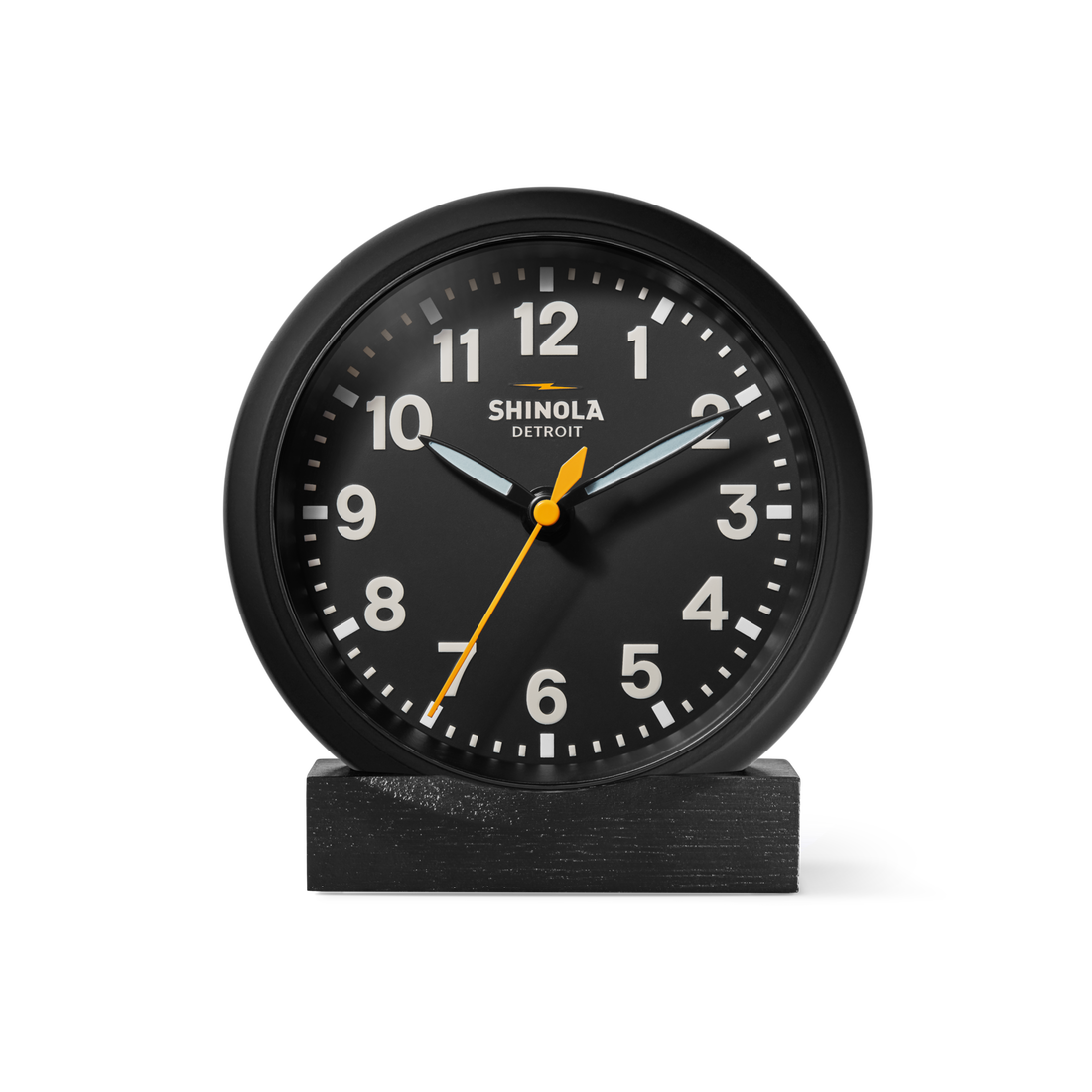 The Shinola 6" Runwell Clock features a black analog face with white numerals, white hour and minute hands, and a yellow second hand. It sits on an oak stand, highlighting Shinola craftsmanship against a white background.
