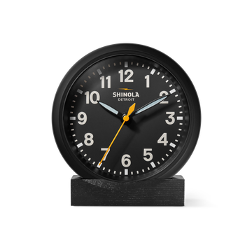 The Shinola 6" Runwell Clock features a black analog face with white numerals, white hour and minute hands, and a yellow second hand. It sits on an oak stand, highlighting Shinola craftsmanship against a white background.