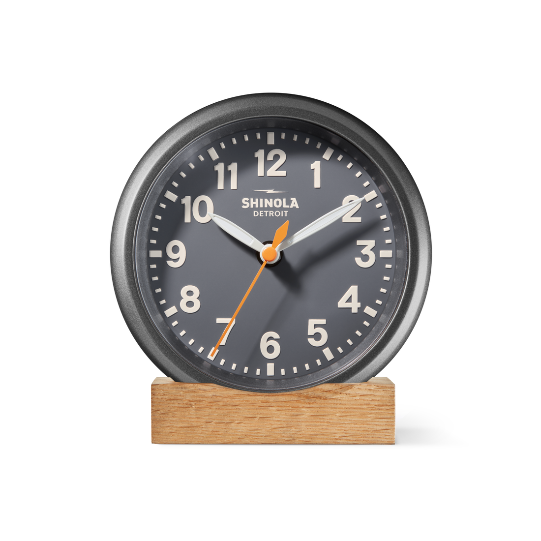 The Shinola 6" Runwell Clock in dark gray, featuring white numbers and hands with an orange second hand, sits on a wooden base. Precision-crafted in Detroit, it displays the brand's name on its face against a white background.