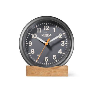 The Shinola 6" Runwell Clock in dark gray, featuring white numbers and hands with an orange second hand, sits on a wooden base. Precision-crafted in Detroit, it displays the brand's name on its face against a white background.